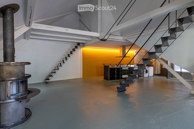 Open-plan space with a modern staircase, kitchen area featuring a central island, oven, and washing machine. Exposed metal chimney and industrial-style lighting.
