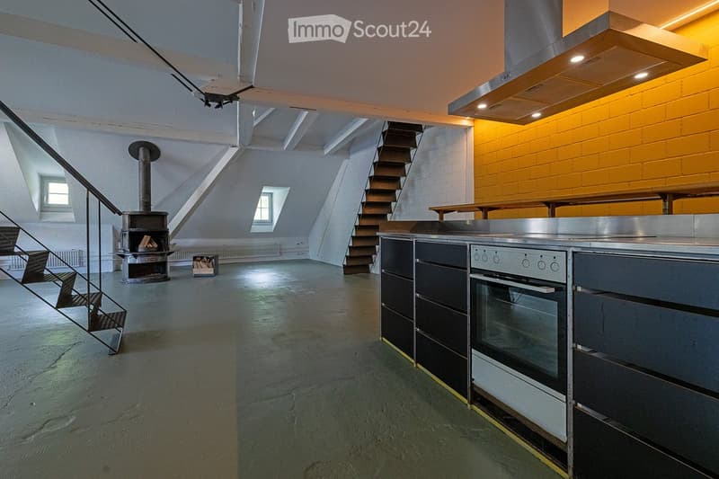 Open-plan space with a modern kitchen area, a staircase leading upwards, and a central fireplace. The kitchen features stainless steel appliances, including an oven and a range hood, with dark cabinets. A wooden table is visible in the background, and the