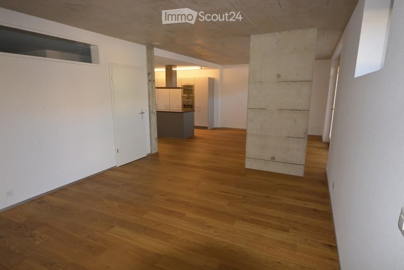 Empty room with wooden flooring, white walls, and a visible kitchen area in the background. No furniture or equipment present.