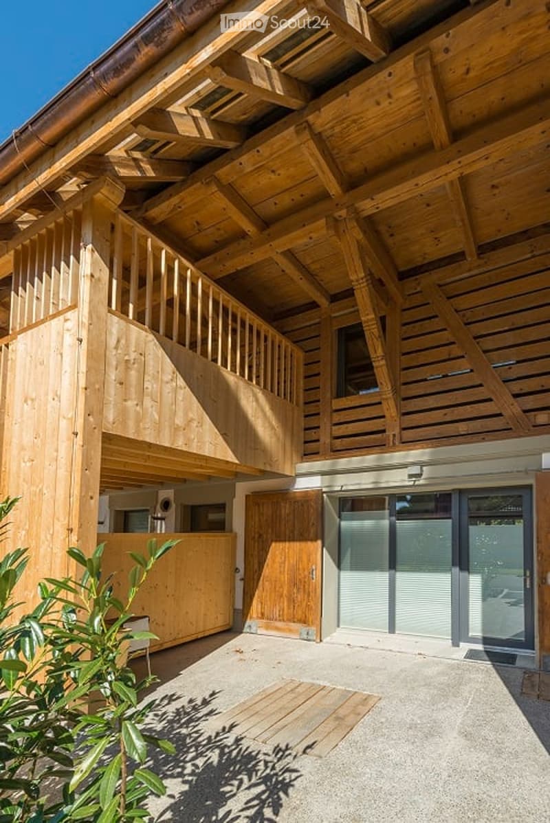 Modern wooden building with a raised ground floor, balcony, and large windows. Features include a wooden staircase, garage entrance, and a small outdoor seating area.