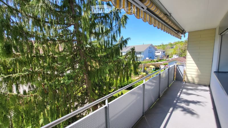 Balcony with metal railing, overlooking a lush garden area with trees and neighboring houses.