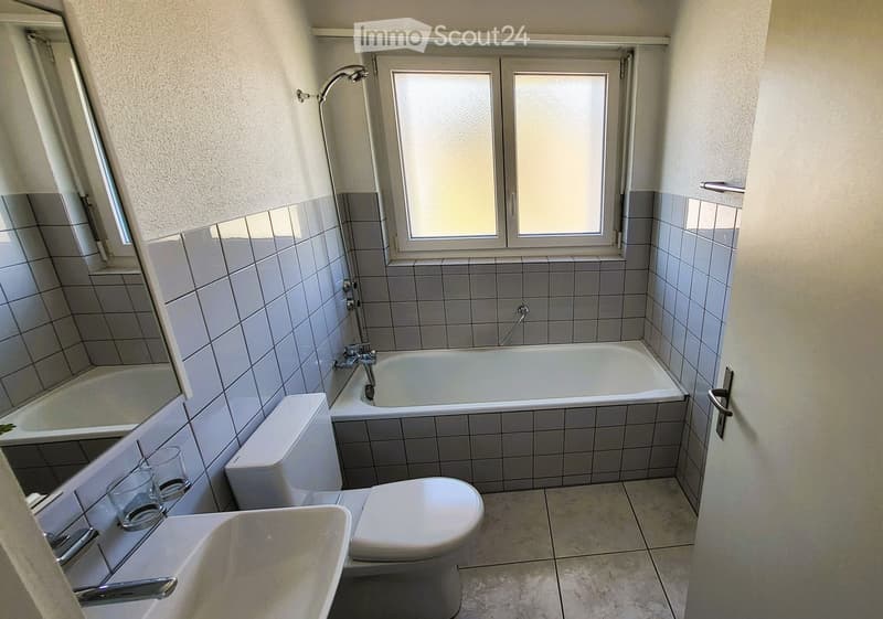 Bathroom with a bathtub, toilet, sink, and showerhead. The walls are tiled, and there is a window above the bathtub.