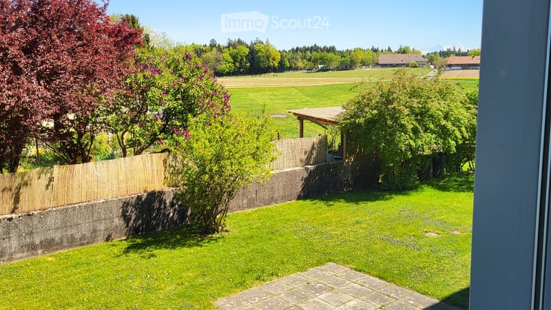 Well-maintained garden with green grass, a wooden fence, and a small wooden pergola. The background features a scenic view of fields, trees, and distant mountains.