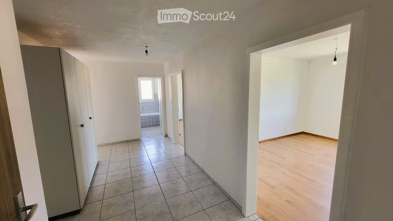 Empty hallway with tiled floor and wooden flooring leading to a small bathroom and another room.