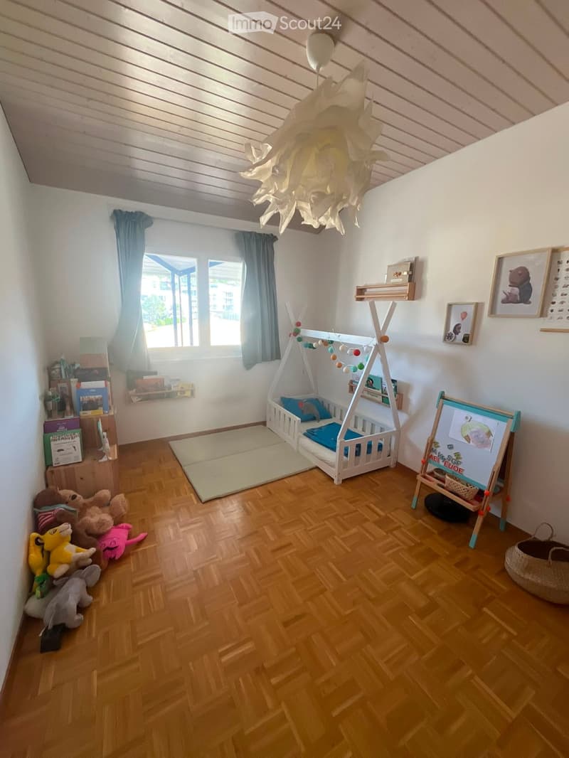 Brightly lit children's bedroom with wooden flooring, a white wooden bed with blue bedding, colorful stuffed animals, a hanging paper lamp, and a small easel with a drawing on it.