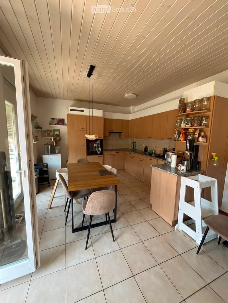 Modern kitchen with light wooden cabinets, granite countertop, dining table with chairs, and various appliances including a refrigerator, microwave, and oven. The space features a kitchen island with a stool and a hanging light fixture.