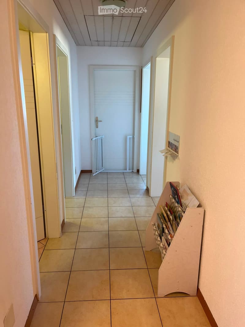 A clean, tiled hallway with white walls, a closed door at the end, and a small shelf holding books and magazines on the right side.