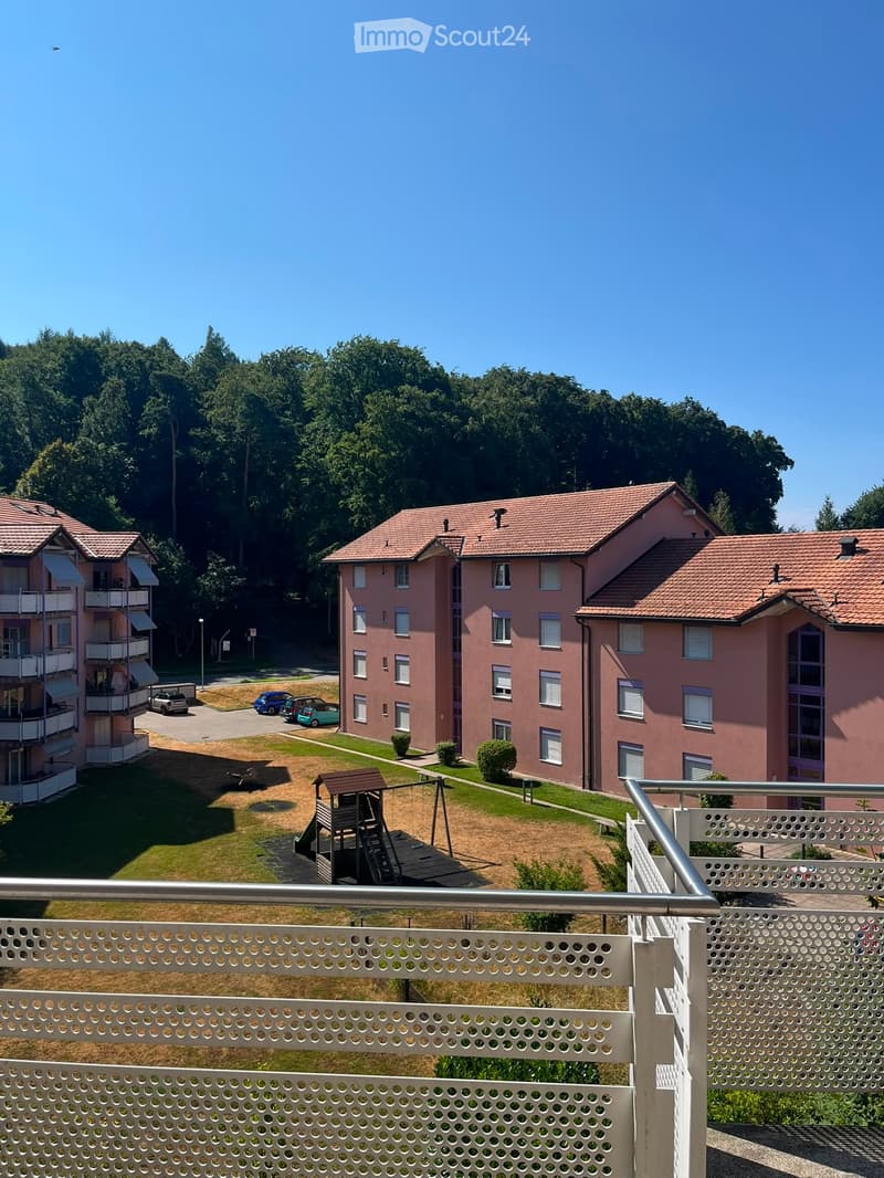 View from a balcony overlooking a residential complex with pink buildings, a playground, and a parking area.