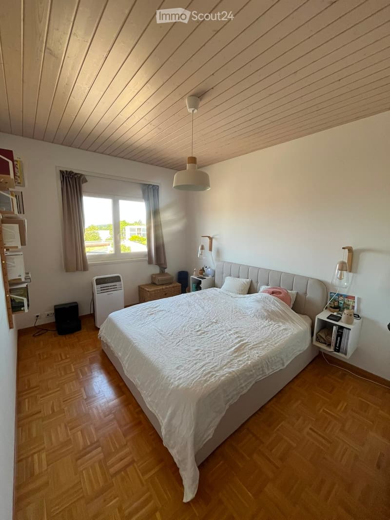 Cozy bedroom with a large bed, wooden flooring, and modern lighting. Features include a nightstand, air conditioner, and shelves with books and decorative items.