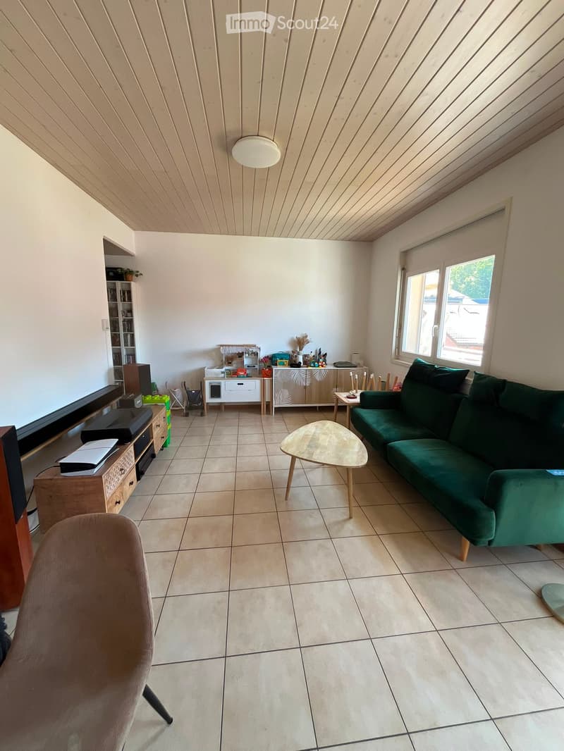 Living room with a green sofa, coffee table, chair, cabinet, and television. The room has tiled flooring and large windows letting in natural light.