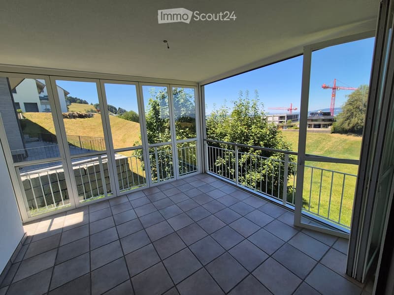 Balcony with tiled floor, large windows, metal railing, overlooking a green landscape with trees and a construction crane in the distance.