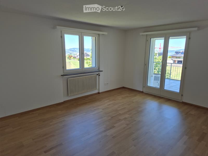 Empty room with wooden flooring, white walls, large windows, and a sliding glass door leading to a balcony.