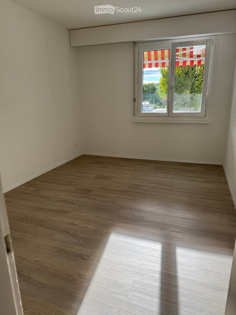 Empty room with wooden flooring and a large window with blinds, showing a view of trees and sky outside.