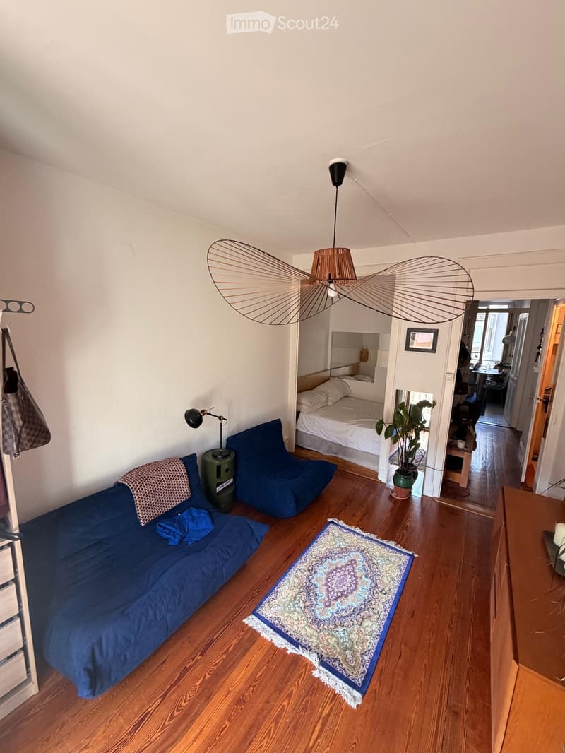 A cozy living space with a blue sofa, a bed in the background, a patterned rug, a hanging lamp, and a small table with a plant.