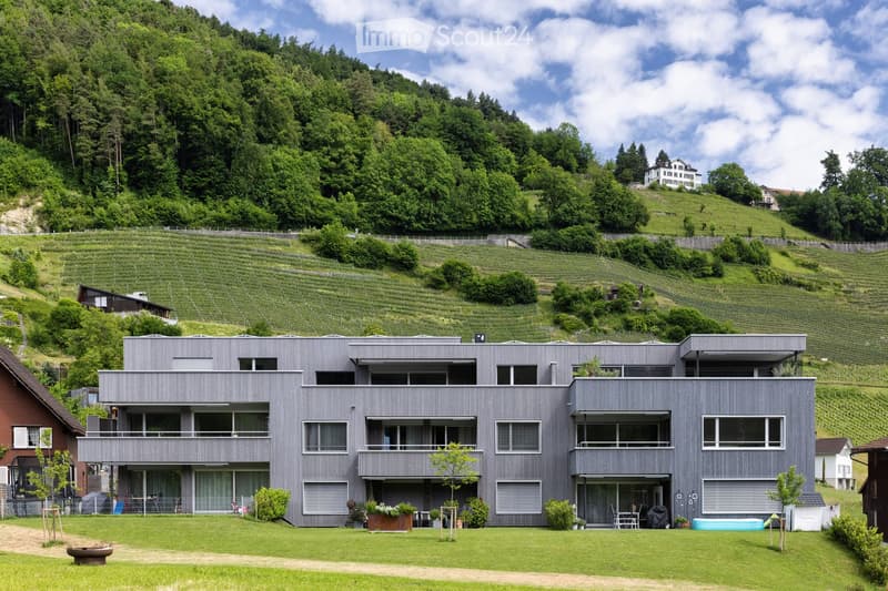 Modern two-story house with grey exterior, large windows, and balconies, surrounded by a well-maintained lawn and vineyard landscape.