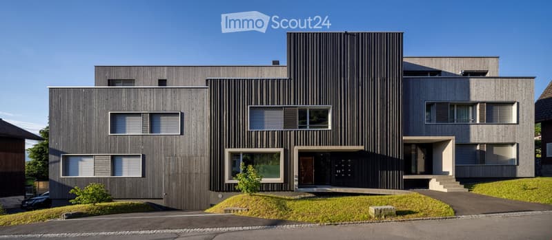 Modern two-story house with dark wood siding, large windows, raised ground floor, and a small landscaped area in front.
