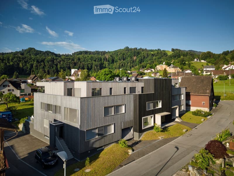 Modern two-story house with grey wooden facade, large windows, and a small garden area.