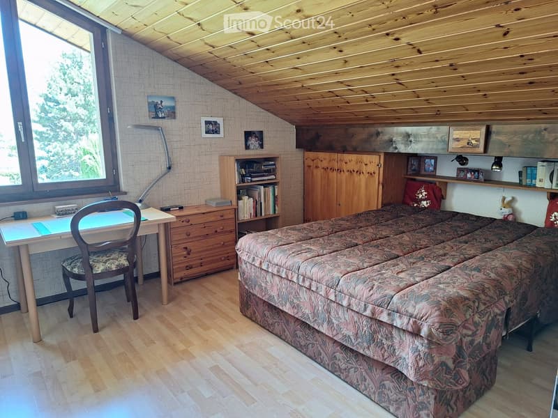Cozy attic bedroom with wooden slanted ceiling, large window, patterned sofa, desk with chair, bookshelf, and cabinet.
