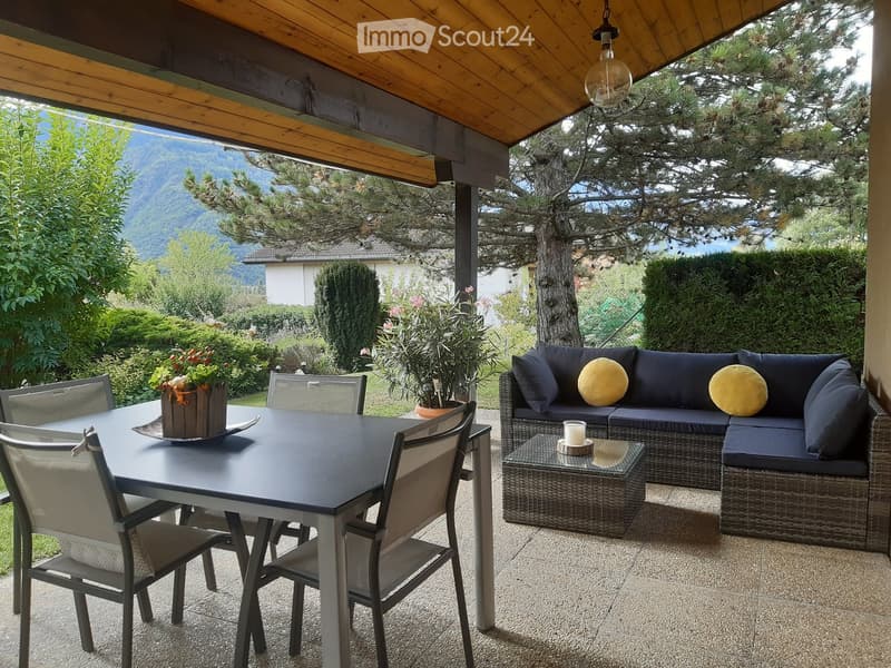 A covered outdoor patio with a wicker sofa, glass coffee table, dining table with chairs, and a hanging light fixture. The view shows a scenic mountain landscape.