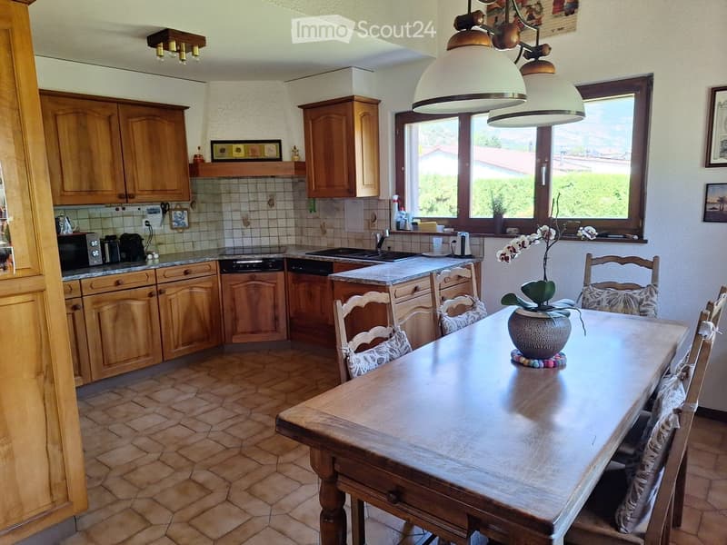 Bright kitchen with wooden cabinets, granite countertops, and a dining area featuring a wooden table and chairs. The space includes a sink, microwave, coffee maker, and a flower pot on the table.
