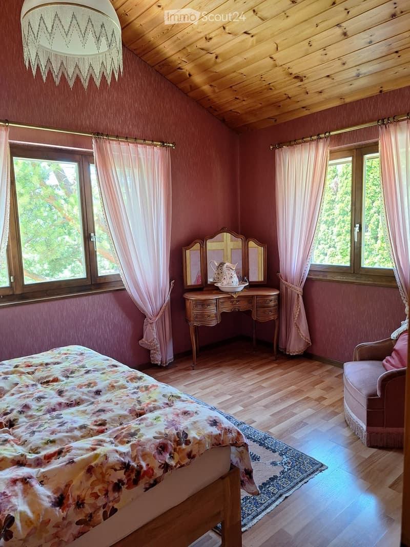 Cozy bedroom with wooden floors, pink walls, large windows with curtains, a floral bedspread, a small sofa, a vanity table with mirrors, and a chandelier.