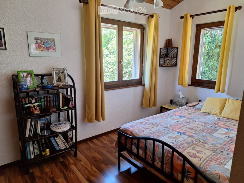Cozy bedroom with wooden flooring, a single bed with colorful bedding, a bookshelf filled with books and decorative items, and large windows with yellow curtains.
