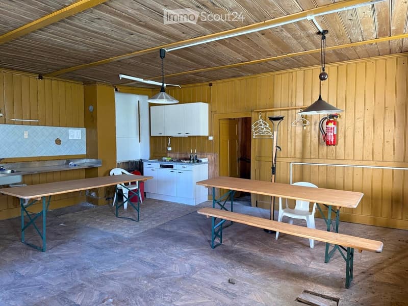 A spacious room with wooden walls and ceiling, featuring two long tables with benches, a kitchen area with cabinets and a sink, and a fire extinguisher mounted on the wall.