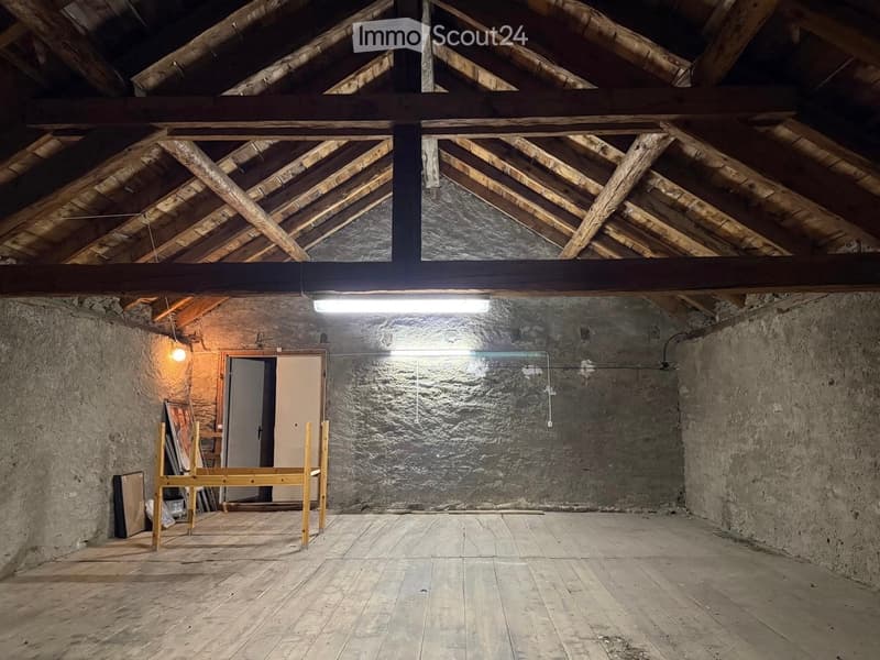 Empty attic space with exposed wooden beams, stone walls, wooden floor, and a single light fixture.