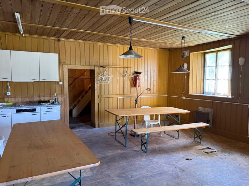 A rustic wooden room with a large wooden table, chairs, a bench, cabinets, and an oven. The room features wooden walls and a wooden ceiling with exposed beams.