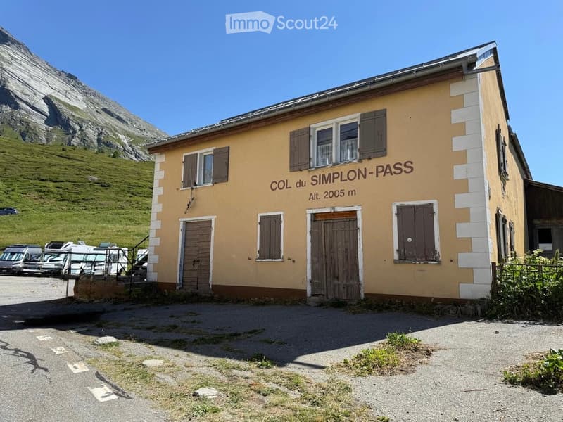 A small, yellow building with brown shutters and a wooden door, labeled "COL du SIMPLON-PASS" with an altitude of 2005 m. It is situated near a parking area with several vehicles and a mountainous backdrop.