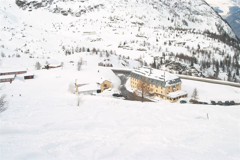 A large, multi-story building surrounded by snow-covered mountains and trees, with several smaller structures and parked cars nearby.