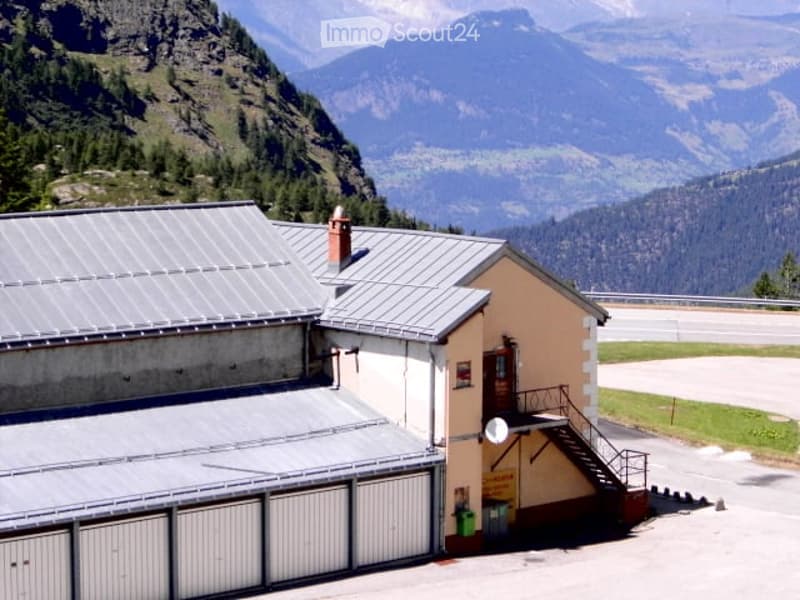 A small building with a grey metal roof and a beige facade, situated in a mountainous area with a scenic view of the surrounding landscape.