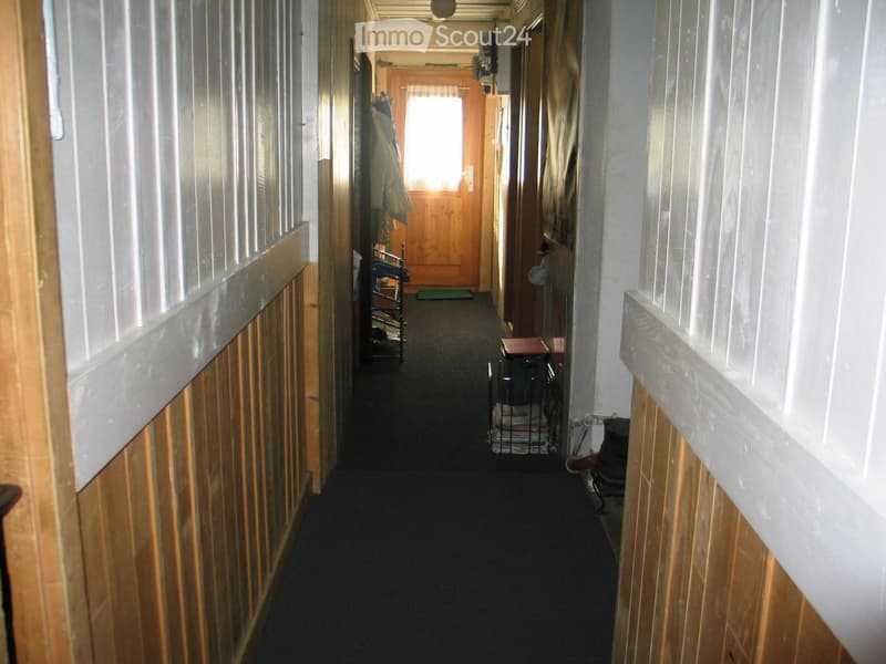 A narrow hallway with wooden panel walls, carpeted floor, and several doors leading to different rooms. A small table and a coat rack are visible on the left side.