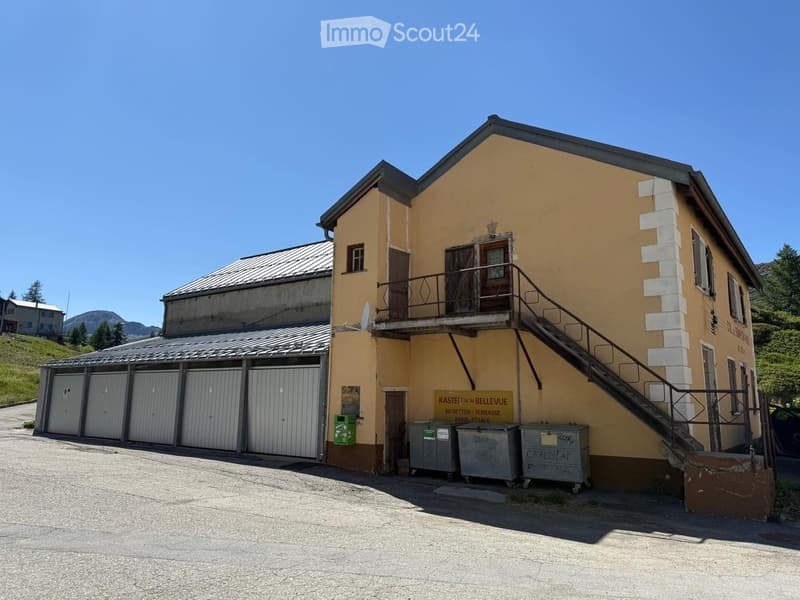 Two-story yellow building with a balcony, garage, and signage reading "Rastei im Bellevue. 80 Betten - Terrasse."