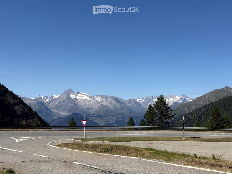 A scenic mountain road with a clear blue sky, snow-capped peaks, and a road sign in the foreground.