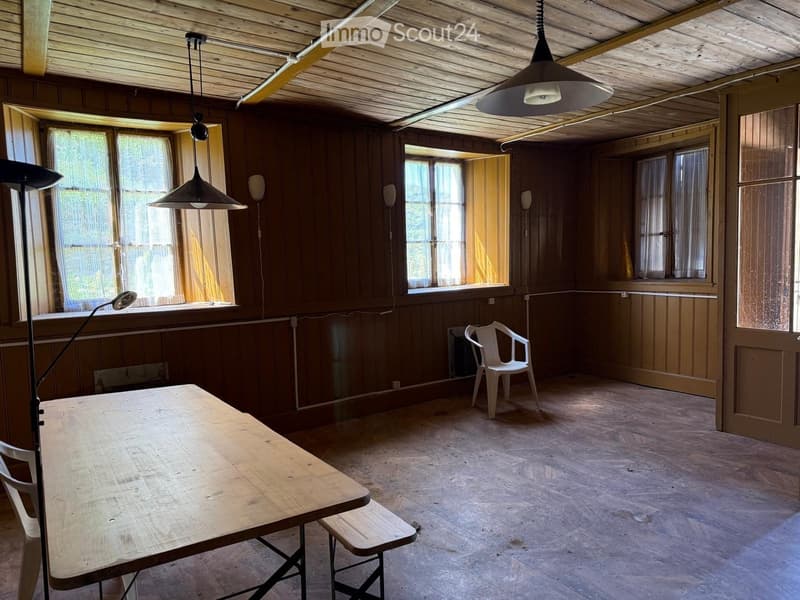 A wooden-paneled room with a large wooden table and a single white plastic chair, natural light from multiple windows, and exposed wooden beams on the ceiling.