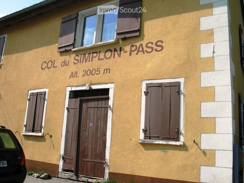 Exterior view of a two-story building with yellow walls, wooden shutters, and a sign reading "COL du SIMPLON-PASS Alt. 2005 m".