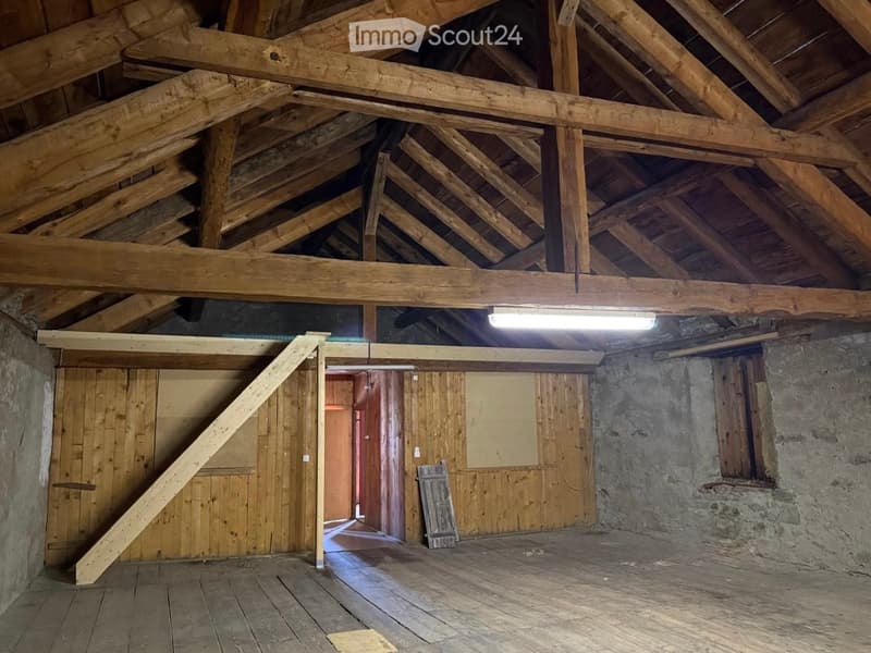 An empty attic space with exposed wooden beams, wooden floor, and stone walls. A single light fixture hangs from the ceiling, and a wooden ladder leads to another room.