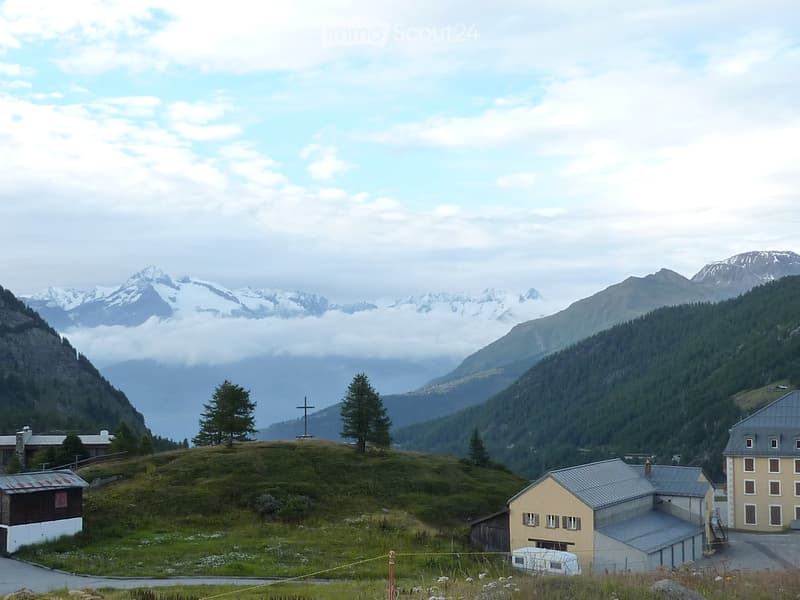 A small cluster of buildings on a hillside with snow-capped mountains in the background, including a cross on a hilltop.