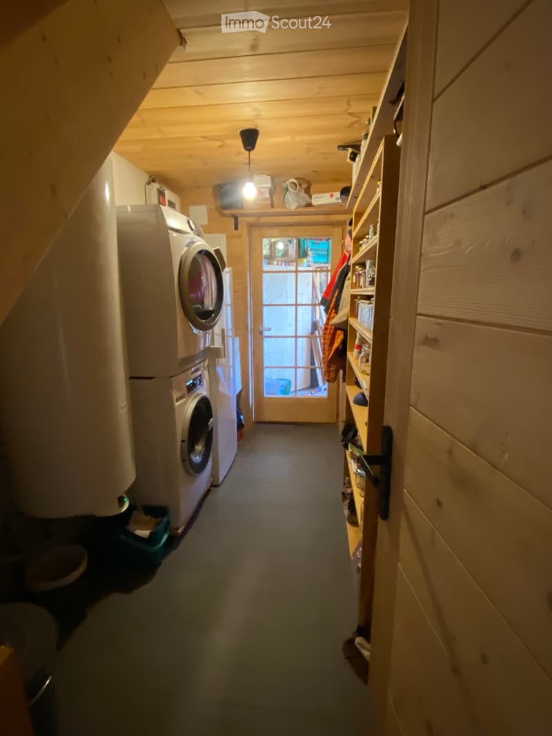 A narrow hallway with wooden walls and ceiling, featuring a washing machine on the left and a shelf with various items on the right, leading to a door with a glass panel.