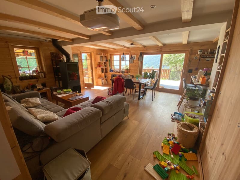Cozy living room with wooden beams, large windows, and a view of the mountains. Features a grey sectional sofa, coffee table, dining area with table and chairs, and a fireplace.