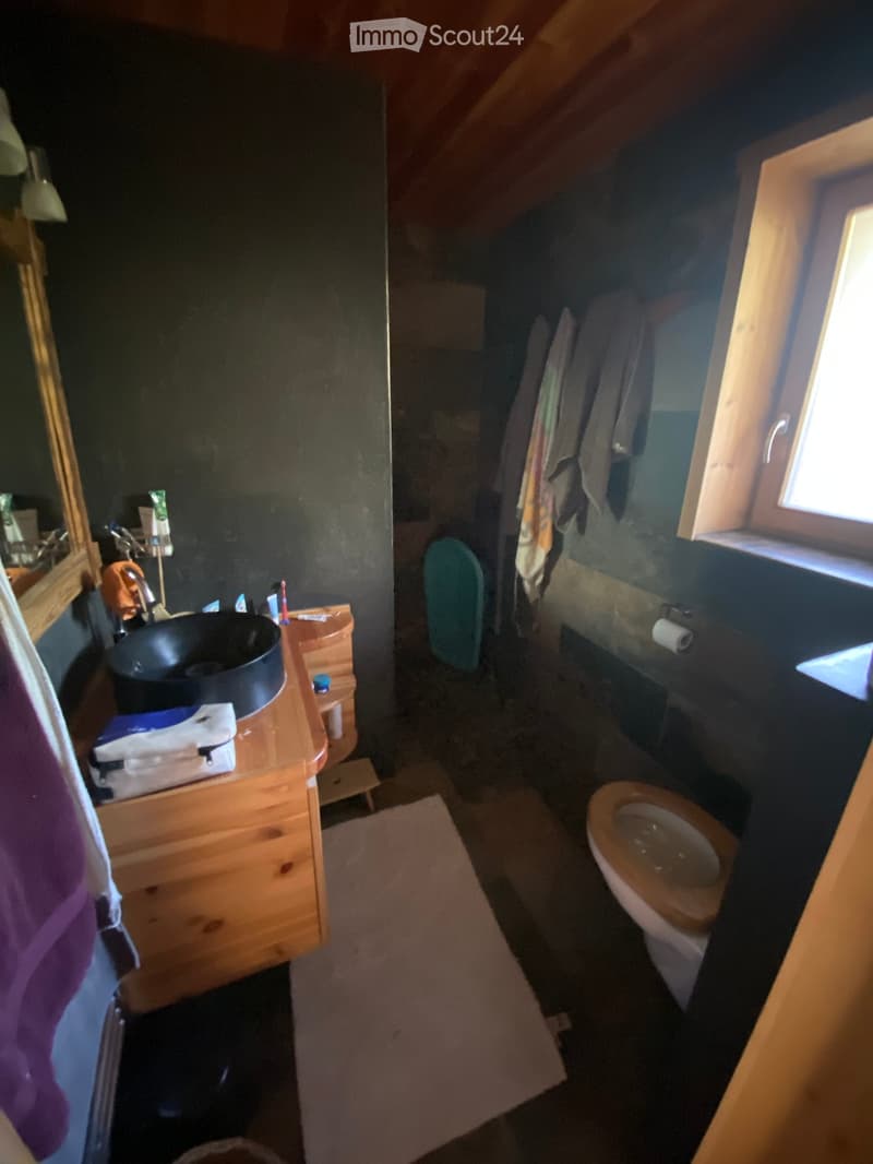 Cozy attic bathroom with wooden sink cabinet, black sink, toilet, mirror, and towels hanging on a rack. Natural light from a window illuminates the space.