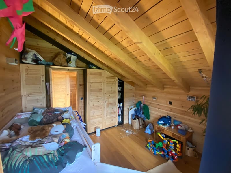 A cozy attic bedroom with wooden walls and slanted ceilings, featuring a bed with colorful bedding, a wardrobe, a bookshelf, and a small table with toys.