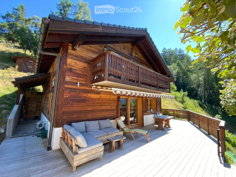 Wooden deck area with a grey sofa, wooden chair, small table, and stool, attached to a wooden house with a balcony and surrounding greenery.