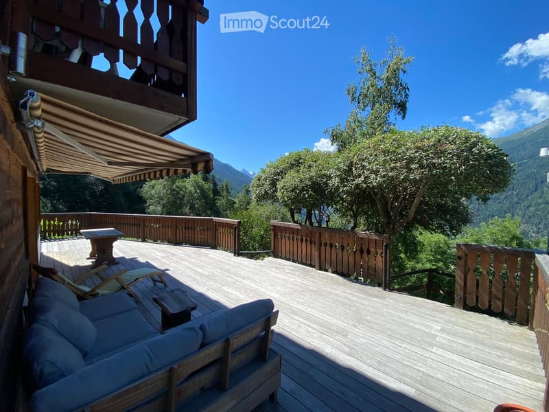 Wooden balcony with a sofa, chairs, and a small table, overlooking a lush green landscape with mountains in the background.