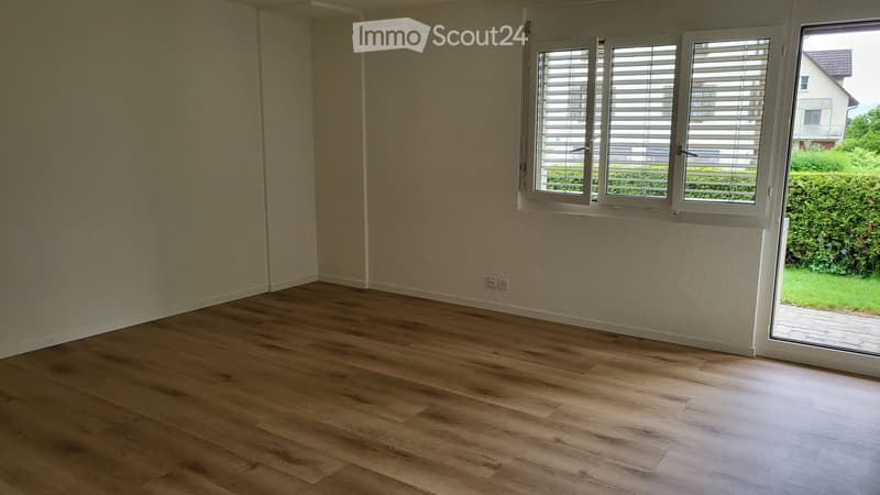 Empty room with wooden flooring and large windows with blinds, showing a view of a garden and neighboring house.