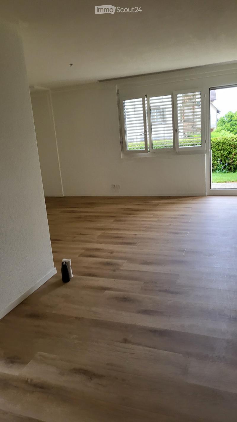 Empty room with wooden flooring, white walls, and large windows with blinds. A small black object is placed near the wall.