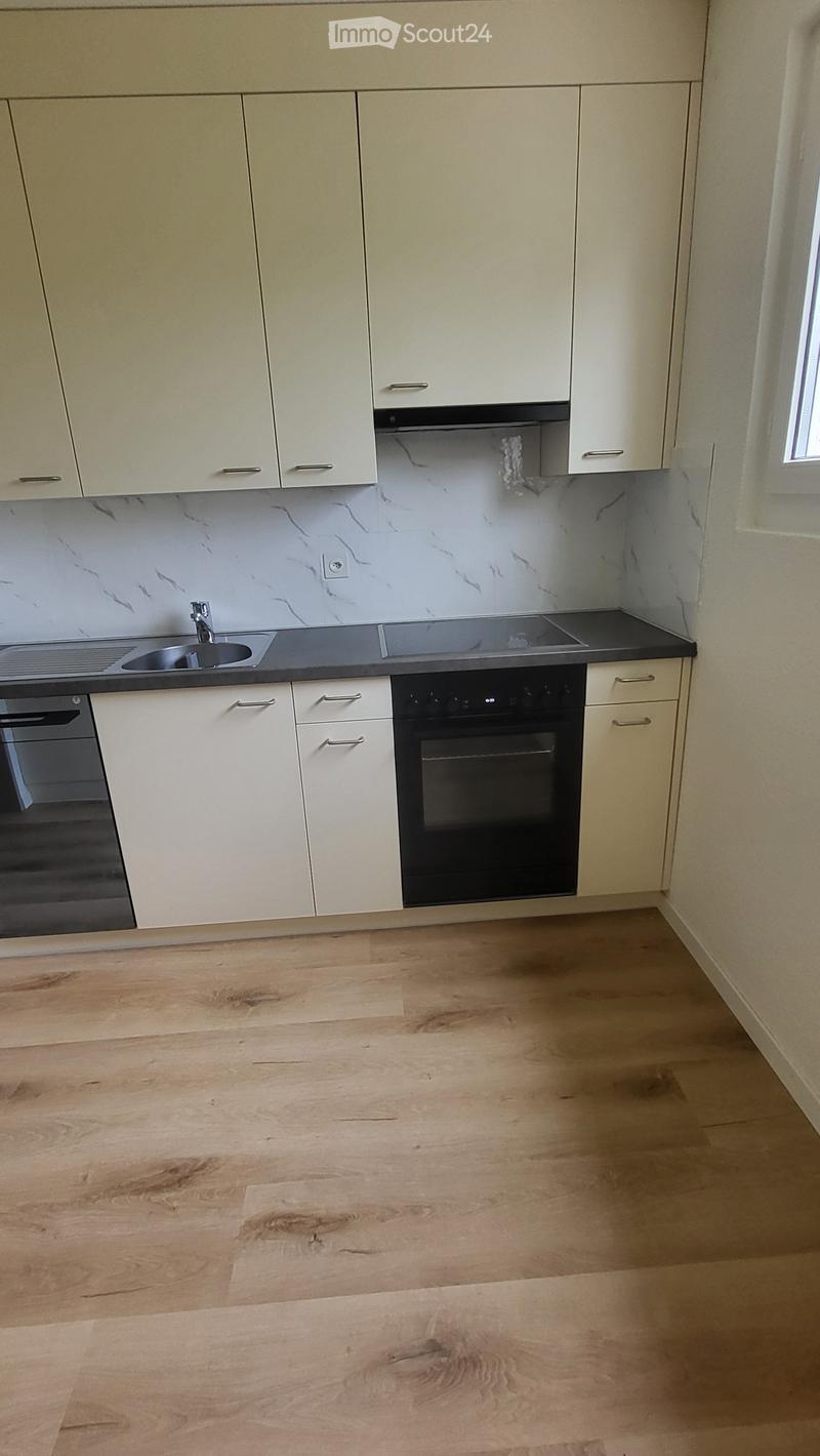 Empty kitchen with light wooden flooring, white cabinets, and built-in appliances including a sink, stove, and oven. The backsplash features a marble-like design.