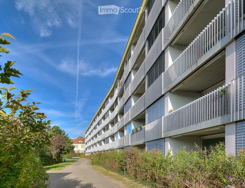 Modern multi-story apartment building with balconies, surrounded by greenery and a clear blue sky.