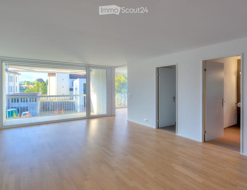 Empty room with light wooden flooring, white walls, large sliding glass doors leading to a balcony, and two open doorways.
